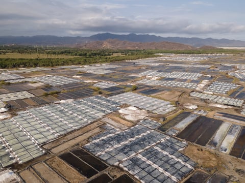 Aerial view of large greenhouse agricultural complex with white and dark plastic-covered structures sprawling across a flat valley beneath mountain ranges