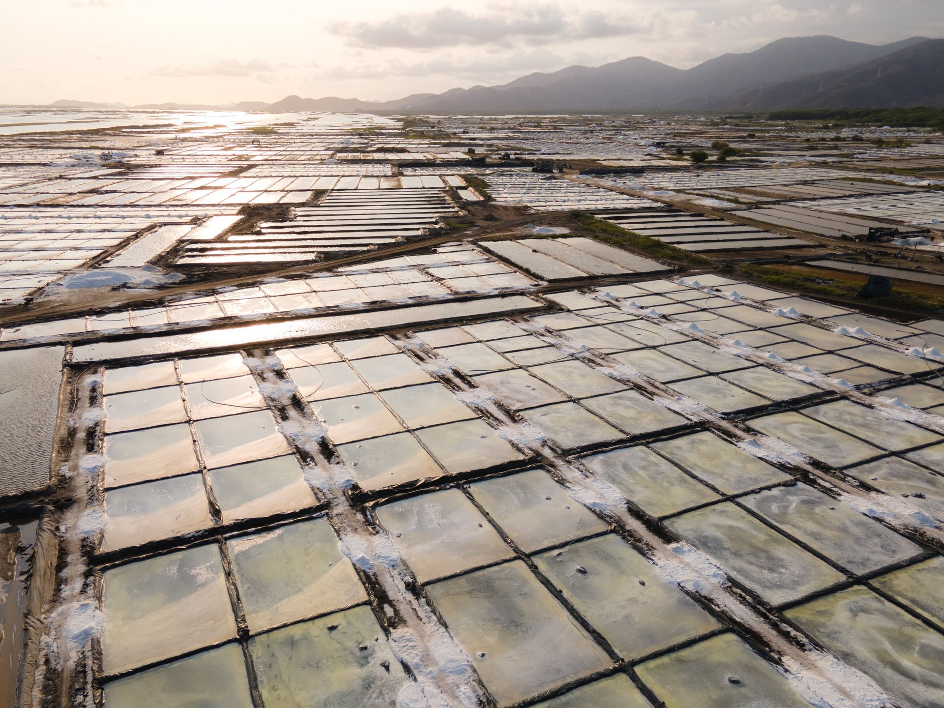 Aerial view of salt ponds with water-filled rectangular pools arranged in rows, mountains visible in distant background under hazy sky